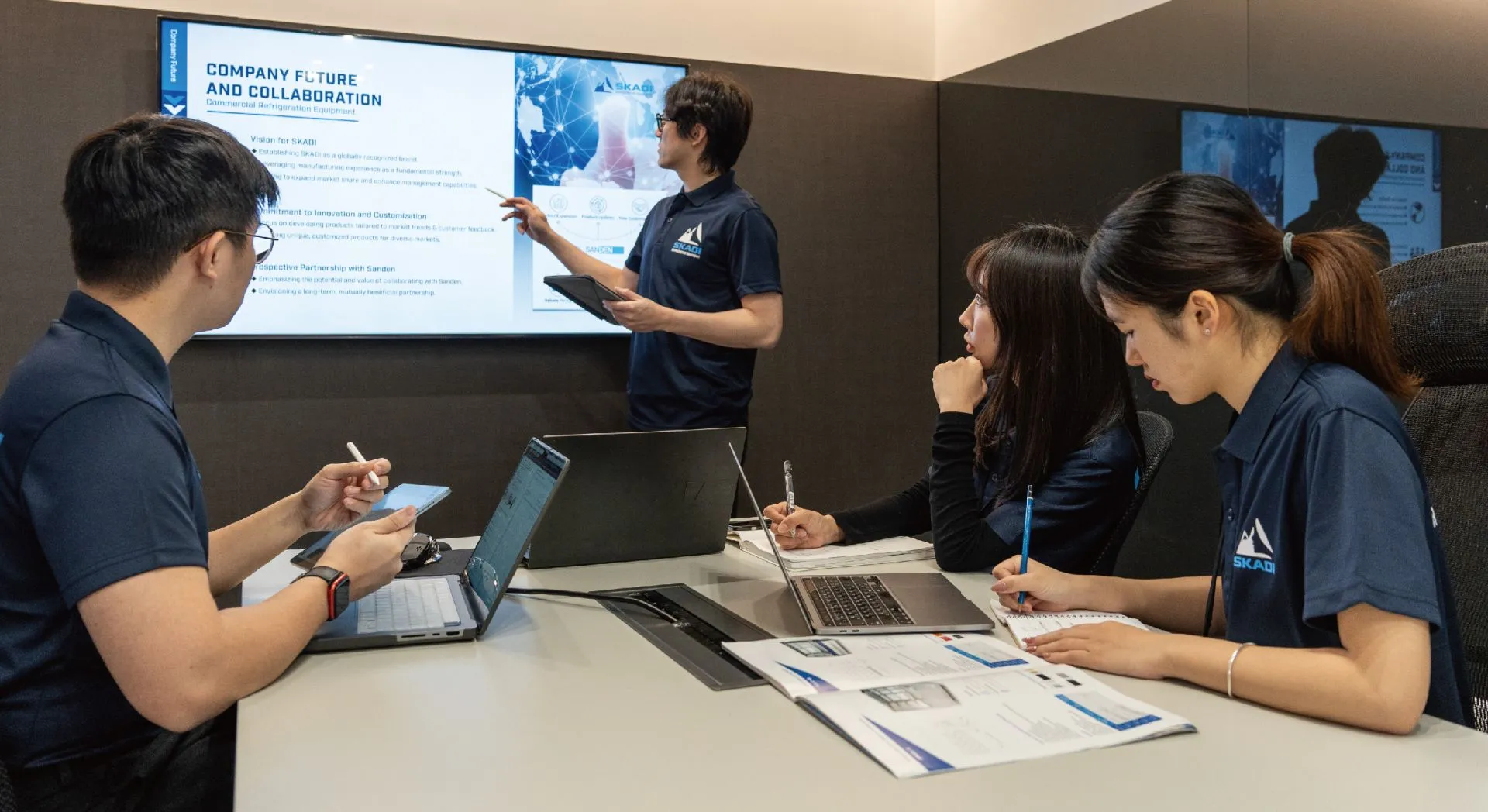 Skadi employees in a meeting room, discussing company goals and quality standards. One employee is standing and pointing to a presentation on a large screen, which outlines the company's vision for future collaboration and innovation in commercial refrigeration equipment. Three other employees are seated at the table with laptops and documents, taking notes and engaging in the discussion. All are wearing Skadi-branded shirts, indicating a professional setting focused on company strategy and product quality.