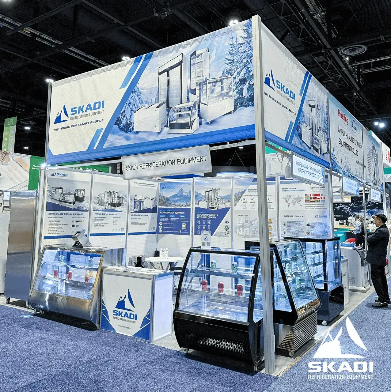 Skadi Refrigeration Equipment booth at the Hotelex exhibition. The booth showcases various refrigerated display cases, with informational posters and banners about Skadi's products, including features and technical specifications. The booth has a large banner with the Skadi logo, featuring images of their refrigeration products against a snowy mountain backdrop. Two display cases in the front hold drinks and packaged items, and a sales representative is seen engaging with attendees.