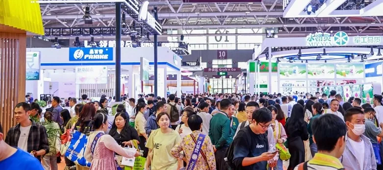 A bustling crowd of attendees at a Hotelex exhibition, walking through various booths and displays in a large convention hall. The image shows people engaging with vendors, many carrying shopping bags and promotional materials. Booths from different brands, such as Panmax, are visible, along with bright signage and lighting. The atmosphere is busy and lively, reflecting a well-attended trade show event.